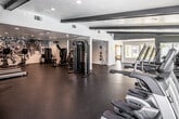 This comprehensive fitness center is thoughtfully organized across a single floor into several distinct workout zones, each designed to support a different aspect of physical training. The cardio area features treadmills, ellipticals, and stair climbers arranged beneath decorative dark ceiling beams, with recessed lighting and windows that bring in natural light and outdoor views. A dedicated indoor cycling studio offers a full row of spinning bikes on warm hardwood flooring, complemented by large wall mirrors and a vibrant, colorful accent wall that energizes the space. The strength and functional training area is generously sized with durable rubber flooring, housing cable machines, dual functional trainers, a dumbbell rack, medicine balls, TRX suspension trainers, and additional cardio equipment such as an air bike and rowing-style machine. High ceilings in portions of this zone add an open, airy feel. A separate free-motion and selectorized machine zone offers additional strength options alongside a bold athletic mural that adds visual character. Throughout the facility, white structural columns provide architectural definition between zones while maintaining an open floor plan. Recessed lighting, large windows, and clean white-and-gray walls create a bright, motivating environment suited to a wide range of fitness pursuits.