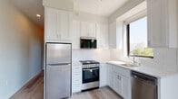 This thoughtfully designed single-floor unit offers a clean, modern aesthetic throughout, with consistent light-toned flooring and neutral wall colors creating a cohesive and welcoming atmosphere.

The kitchen is a standout feature, outfitted with crisp white shaker-style cabinetry extending floor to ceiling for ample storage. A full suite of stainless steel appliances includes a refrigerator, a gas range with oven, an over-the-range microwave, and a dishwasher. A white tile backsplash and a double basin sink complement the bright countertops, while a large window invites natural light and outdoor views, enhancing the overall sense of openness.

The living and sleeping areas continue the light-filled theme, with large windows framing views of surrounding greenery and neighboring rooftops. One room features light wood-style flooring and a wall-mounted HVAC unit, while a second room is finished with plush carpeting and a generous window that floods the space with sunlight. A barn-style sliding door adds a modern design touch.

The bathroom is clean and functional, featuring wood-look tile flooring, a vanity with storage, a toilet, and a small window for natural ventilation. A stacked washer and dryer unit is conveniently tucked within the unit, adding practical in-home laundry access. Overall, the space presents a bright, move-in-ready environment with modern finishes throughout.