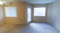 This single-floor residence offers a clean and functional layout designed for comfortable everyday living. The living area serves as the heart of the space, finished with soft, plush wall-to-wall carpeting in a neutral gray tone that adds warmth underfoot. The walls are painted in a light beige and gray palette, creating a calm and cohesive aesthetic throughout.

Natural light is a defining feature of this space, with multiple windows dressed in white vertical blinds that allow for flexible light control while framing views of the surrounding outdoor greenery. A glass-paneled exterior door provides both an additional source of natural light and direct outdoor access, enhancing the sense of openness.

A built-in wall heater unit is conveniently positioned at the base of one wall, ensuring efficient climate control within the living area. The room's simple, uncluttered design offers a versatile canvas for a variety of furnishing arrangements and interior styles.

Overall, the space presents a practical and welcoming environment, with thoughtful details such as consistent neutral finishes and well-placed windows that contribute to a bright, airy atmosphere throughout the single-floor layout.