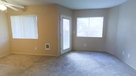 This single-floor residence offers a clean and functional layout designed for comfortable everyday living. The living area serves as the heart of the space, finished with soft, plush wall-to-wall carpeting in a neutral gray tone that adds warmth underfoot. The walls are painted in a light beige and gray palette, creating a calm and cohesive aesthetic throughout.

Natural light is a defining feature of this space, with multiple windows dressed in white vertical blinds that allow for flexible light control while framing views of the surrounding outdoor greenery. A glass-paneled exterior door provides both an additional source of natural light and direct outdoor access, enhancing the sense of openness.

A built-in wall heater unit is conveniently positioned at the base of one wall, ensuring efficient climate control within the living area. The room's simple, uncluttered design offers a versatile canvas for a variety of furnishing arrangements and interior styles.

Overall, the space presents a practical and welcoming environment, with thoughtful details such as consistent neutral finishes and well-placed windows that contribute to a bright, airy atmosphere throughout the single-floor layout.