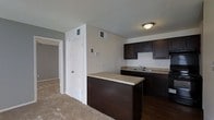This single-story space showcases a well-appointed kitchen as its centerpiece, designed with rich, dark espresso-toned cabinetry that extends from base to upper units, creating a bold contrast against the neutral beige walls. The countertops feature a light granite-style surface, providing a bright counterbalance to the dark cabinetry throughout. A kitchen island with matching dark base and granite-style top offers additional workspace and serves as a natural dividing point between the kitchen and adjacent living areas. The kitchen is equipped with a stainless steel sink set into the perimeter countertop, with ample upper and lower cabinet storage. The flooring transitions from dark hardwood-style material in the kitchen to plush carpeting in the adjoining spaces, delineating the functional zones of the layout. Recessed vents are positioned throughout the ceiling and lower walls, indicating a central HVAC system. Overhead lighting provides consistent illumination. A doorway visible from the kitchen leads to additional rooms, suggesting a practical, connected floor plan. The textured ceiling and clean wall finishes contribute to a straightforward, functional aesthetic throughout.