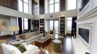 This distinguished single-floor residence showcases exceptional architectural design centered around a dramatic double-height living area with soaring ceilings that create an immediate sense of grandeur. Towering floor-to-ceiling windows, framed in rich dark wood, flood the interior with abundant natural light while offering sweeping panoramic views of the surrounding landscape through a private balcony access point.

The main living area is anchored by a refined arrangement of furnishings, including a tufted velvet sofa, a luxurious leather ottoman serving as a central coffee table, and an assortment of elegant accent chairs. A boldly patterned area rug grounds the seating arrangement atop gleaming dark hardwood floors that extend throughout.

Built-in bookcases flanking the windows provide both architectural symmetry and functional display space, contributing to the space's sophisticated, library-inspired character. Track lighting installed along the upper walls complements the natural light and highlights the room's vertical scale.

Additional seating and reading areas are thoughtfully arranged near the windows, taking advantage of the remarkable views and natural illumination. The overall ambiance blends classic elegance with a warm, livable sensibility — making this a space that is as inviting as it is architecturally impressive.