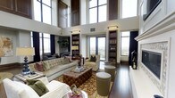 This distinguished single-floor residence showcases exceptional architectural design centered around a dramatic double-height living area with soaring ceilings that create an immediate sense of grandeur. Towering floor-to-ceiling windows, framed in rich dark wood, flood the interior with abundant natural light while offering sweeping panoramic views of the surrounding landscape through a private balcony access point.

The main living area is anchored by a refined arrangement of furnishings, including a tufted velvet sofa, a luxurious leather ottoman serving as a central coffee table, and an assortment of elegant accent chairs. A boldly patterned area rug grounds the seating arrangement atop gleaming dark hardwood floors that extend throughout.

Built-in bookcases flanking the windows provide both architectural symmetry and functional display space, contributing to the space's sophisticated, library-inspired character. Track lighting installed along the upper walls complements the natural light and highlights the room's vertical scale.

Additional seating and reading areas are thoughtfully arranged near the windows, taking advantage of the remarkable views and natural illumination. The overall ambiance blends classic elegance with a warm, livable sensibility — making this a space that is as inviting as it is architecturally impressive.