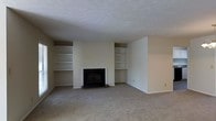 This single-floor space offers a thoughtfully designed living area that serves as the heart of the home. The room is anchored by a classic fireplace with a sleek black surround and hearth, flanked symmetrically on both sides by built-in open shelving units. These shelves provide generous display and storage space, adding both functionality and architectural character to the room. The walls are finished in a soft, neutral tone that creates a calm and inviting atmosphere, while the textured popcorn ceiling adds a layer of visual interest overhead. Wall-to-wall carpeting in a warm, neutral hue runs throughout the space, contributing to a comfortable and understated aesthetic. The open layout of the living area allows for flexible furniture arrangement, accommodating a variety of living and entertaining configurations. The overall design is clean and straightforward, with an emphasis on practicality and comfort. The neutral palette throughout makes the space highly adaptable, offering a versatile canvas for personalization with furnishings and décor. This is a well-structured, move-in-ready space ideal for those seeking a quiet and functional single-level living environment.