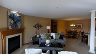This single-floor residence offers an open-concept layout that seamlessly blends comfort and style. The living room serves as the heart of the space, featuring warm tan-hued walls that create an inviting atmosphere. A gray upholstered sofa, accented with decorative pillows in navy, green, and floral patterns, anchors the seating area around a dark wood coffee table. A classic fireplace mantel in warm wood tones adds a traditional focal point to the room, while a striking black wrought-iron wall medallion adds an artistic touch. Soft carpeting underfoot enhances the cozy ambiance throughout. The living area flows naturally into the dining space, which is illuminated by a hanging chandelier that casts a warm glow over the dining table and chair set. Neutral carpeting and consistent warm wall tones create a cohesive visual connection between the two areas. A hallway visible from the main living space suggests additional rooms beyond, completing this well-appointed single-level home. The overall design balances functional living with thoughtful decorative details, resulting in a space that feels both comfortable and refined.