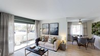 This single-floor residence offers a welcoming and well-appointed interior designed for comfortable everyday living. The open-concept layout seamlessly connects the living and dining areas, creating a sense of spaciousness and easy flow throughout. Light wood-tone flooring runs throughout the main living space, complementing the neutral, contemporary color palette.

The living area is anchored by a stylish arrangement featuring a plush sofa adorned with patterned accent pillows, a geometric-patterned area rug, and a sleek coffee table. Two large framed art pieces hang above the sofa, adding a sophisticated, modern touch to the wall. A brass arc floor lamp provides warm ambient lighting, enhancing the inviting atmosphere.

A striking feature of the living room is the large sliding glass door that opens to an outdoor balcony or patio, flooding the space with natural light and offering pleasant views of surrounding greenery and trees. Soft linen-toned curtains frame both the sliding door and the additional window, adding an elegant finishing detail.

Adjacent to the living area, the dining space is visible and equally well-lit, featuring a compact dining table with chairs beneath a ceiling fan. The cohesive design throughout creates a bright, airy, and functional living environment with a clean, modern aesthetic.