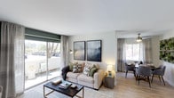 This single-floor residence offers a welcoming and well-appointed interior designed for comfortable everyday living. The open-concept layout seamlessly connects the living and dining areas, creating a sense of spaciousness and easy flow throughout. Light wood-tone flooring runs throughout the main living space, complementing the neutral, contemporary color palette.

The living area is anchored by a stylish arrangement featuring a plush sofa adorned with patterned accent pillows, a geometric-patterned area rug, and a sleek coffee table. Two large framed art pieces hang above the sofa, adding a sophisticated, modern touch to the wall. A brass arc floor lamp provides warm ambient lighting, enhancing the inviting atmosphere.

A striking feature of the living room is the large sliding glass door that opens to an outdoor balcony or patio, flooding the space with natural light and offering pleasant views of surrounding greenery and trees. Soft linen-toned curtains frame both the sliding door and the additional window, adding an elegant finishing detail.

Adjacent to the living area, the dining space is visible and equally well-lit, featuring a compact dining table with chairs beneath a ceiling fan. The cohesive design throughout creates a bright, airy, and functional living environment with a clean, modern aesthetic.
