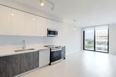 Located at 150 S Bryan Rd in Dania Beach, Florida, this modern residence offers a thoughtfully designed one-bedroom, two-bathroom layout across a single floor. The open-concept living and kitchen area is anchored by a sleek, contemporary kitchen featuring two-tone cabinetry — warm wood-grain lower cabinets paired with crisp white upper cabinets — complemented by white quartz countertops, a white subway tile backsplash, and a full suite of stainless steel appliances including a French door refrigerator, electric range, and over-the-range microwave. Light wood-toned flooring flows throughout, enhancing the bright, airy atmosphere. The living area opens to large floor-to-ceiling sliding glass doors leading to a balcony, flooding the space with natural light and providing outdoor views. The bedroom is a calm, neutral retreat with a large window framing views of the surrounding community and ample room for furnishings. The two bathrooms are finished in a clean, modern style, one featuring a soaking tub and shower combination with light stone-look tile surround, a floating wood vanity, and contemporary fixtures. Track lighting and pendant accents throughout add a polished, functional touch to this well-appointed Dania Beach residence.