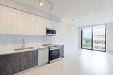 Located at 150 S Bryan Rd in Dania Beach, Florida, this modern residence offers a thoughtfully designed one-bedroom, two-bathroom layout across a single floor. The open-concept living and kitchen area is anchored by a sleek, contemporary kitchen featuring two-tone cabinetry — warm wood-grain lower cabinets paired with crisp white upper cabinets — complemented by white quartz countertops, a white subway tile backsplash, and a full suite of stainless steel appliances including a French door refrigerator, electric range, and over-the-range microwave. Light wood-toned flooring flows throughout, enhancing the bright, airy atmosphere. The living area opens to large floor-to-ceiling sliding glass doors leading to a balcony, flooding the space with natural light and providing outdoor views. The bedroom is a calm, neutral retreat with a large window framing views of the surrounding community and ample room for furnishings. The two bathrooms are finished in a clean, modern style, one featuring a soaking tub and shower combination with light stone-look tile surround, a floating wood vanity, and contemporary fixtures. Track lighting and pendant accents throughout add a polished, functional touch to this well-appointed Dania Beach residence.