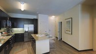 This well-appointed single-floor residence showcases a thoughtfully designed open-concept layout that seamlessly connects the kitchen and living areas. The kitchen is a standout feature, highlighted by a large central island with striking granite countertops and seating for casual dining. Dark espresso cabinetry extends to the ceiling, providing generous storage, while stainless steel appliances — including a French door refrigerator with an in-door ice and water dispenser — add a sleek, modern touch. Warm hardwood-style flooring runs throughout the space, lending a cohesive and inviting feel underfoot. The neutral, light-toned walls create a bright and airy ambiance that complements the bold contrast of the dark cabinetry. A white paneled entry door serves as a clean architectural focal point, while an adjacent hallway leads to additional rooms beyond. Subtle recessed and flush-mount lighting provides consistent illumination throughout. Decorative touches, such as a framed landscape print on the wall, add warmth and personality to the space. The overall design balances contemporary style with practical functionality, making it a comfortable and attractive living environment.
