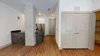This single-floor unit showcases a contemporary design highlighted by warm wood-look flooring that runs throughout the open-concept living and kitchen areas. A distinctive concrete-finish structural column serves as an architectural focal point, visually separating the kitchen from the living space while adding an industrial character to the overall aesthetic.

The kitchen is well-appointed with stainless steel appliances, including a full-size refrigerator, over-the-range microwave, and electric range. White shaker-style cabinetry extends to the upper walls, providing ample storage, while a sleek dark countertop with a subtle sheen offers generous prep space. A light blue tile backsplash adds a refined accent behind the cooking area.

The living area is generously proportioned, featuring large floor-to-ceiling windows and a glass-panel entry door that invite abundant natural light and views of the surrounding greenery. Two ceiling fans with integrated lighting ensure comfortable airflow throughout the space.

The bathroom features white shaker-style cabinetry paired with a dark stone countertop, an undermount sink, and brushed nickel hardware and fixtures, contributing to the unit's cohesive modern aesthetic. Recessed lighting and neutral white walls complete the clean, contemporary look across the entire space.