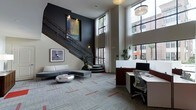 This contemporary space makes an immediate impression with its dramatic double-height ceilings and an elegant staircase adorned with intricate wrought-iron railings, serving as a striking architectural centerpiece. The staircase rises against a deep charcoal-toned accent wall, where carefully positioned artwork adds visual interest and depth.

The lounge area features a streamlined gray sectional sofa paired with a sculptural metallic coffee table, all set atop a patterned carpet with subtle red accents that ground the open layout. Adjacent to the lounge, a dedicated workspace includes a clean-lined white desk station equipped with a monitor and ergonomic seating, ideal for productivity.

Floor-to-ceiling windows dressed with sheer white curtains flood the space with natural light while offering views of lush greenery and surrounding architecture. A pendant light fixture overhead provides warm, ambient illumination. Decorative potted plants are thoughtfully placed to introduce organic texture. The overall ambiance is polished and modern, balancing professional functionality with stylish comfort.