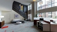 This contemporary space makes an immediate impression with its dramatic double-height ceilings and an elegant staircase adorned with intricate wrought-iron railings, serving as a striking architectural centerpiece. The staircase rises against a deep charcoal-toned accent wall, where carefully positioned artwork adds visual interest and depth.

The lounge area features a streamlined gray sectional sofa paired with a sculptural metallic coffee table, all set atop a patterned carpet with subtle red accents that ground the open layout. Adjacent to the lounge, a dedicated workspace includes a clean-lined white desk station equipped with a monitor and ergonomic seating, ideal for productivity.

Floor-to-ceiling windows dressed with sheer white curtains flood the space with natural light while offering views of lush greenery and surrounding architecture. A pendant light fixture overhead provides warm, ambient illumination. Decorative potted plants are thoughtfully placed to introduce organic texture. The overall ambiance is polished and modern, balancing professional functionality with stylish comfort.