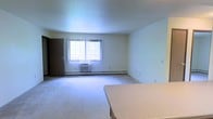 This single-floor residential unit offers a straightforward and functional layout designed for comfortable everyday living. The main living area is finished with wall-to-wall carpeting that adds warmth underfoot and contributes to a quiet, cozy atmosphere throughout the space. Neutral-toned walls and a smooth ceiling keep the interior feeling bright and clean, providing a versatile backdrop for a variety of furnishing styles. A large window fitted with vertical blinds allows natural light to filter into the room, creating an airy feel while maintaining privacy. Beneath the window, a wall-mounted air conditioning unit ensures cooling during warmer months, while baseboard heating running along the perimeter of the room provides reliable warmth in cooler seasons. A standard interior door, finished in a medium wood tone, is visible along one wall, connecting to an adjacent space. Electrical outlets are conveniently positioned along the walls to accommodate modern living needs. The room offers generous open floor space, making it well-suited for flexible furniture arrangements. Overall, the unit presents a practical and livable environment with essential climate control features already in place.