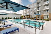 Located at 6101 Richmond Avenue in Houston, Texas, this outdoor amenity space is designed to deliver a resort-style experience within a modern residential community. At its centerpiece is a generously sized lap pool with clearly marked lane dividers, offering crystal-clear water ideal for both recreational swimming and exercise. The pool deck is finished in smooth concrete and provides ample space for lounging, with multiple navy blue chaise lounge chairs positioned along the pool's perimeter for relaxation.

Shade is thoughtfully provided through bold blue cantilever and standard market umbrellas stationed at various points around the deck. A built-in outdoor grilling station is conveniently located nearby, making the space well-suited for outdoor gatherings and social activities.

The courtyard is enclosed by the multi-story building facades, which feature a contemporary mix of brick and light-colored siding with large windows and private balconies overlooking the pool area. Manicured landscaping with shrubs, ornamental grasses, mulched garden beds, and young trees lines the perimeter, adding natural texture and greenery to the hardscaped environment. Gated access points and perimeter fencing contribute to a sense of security and privacy, while open sightlines throughout the space maintain an airy, welcoming atmosphere.