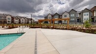Located in Herriman, Utah, this outdoor community amenity space is designed to serve as a central gathering hub for residents. At its heart lies a large, sparkling swimming pool with crystal-clear water, surrounded by a generous concrete pool deck lined with white lounge chairs ideal for sunbathing. Multiple shade structures, including tensile canopy sails and a traditional wooden pergola, provide covered retreat areas throughout the space.

Adjacent to the pool deck is a well-maintained artificial turf lawn, offering additional seating with white lounge chairs positioned beneath the shade canopies. Natural boulder landscaping and mulched planting beds border the lawn, adding texture and a connection to the surrounding Utah terrain. Picnic-style tables are scattered across the hardscaped patio areas, creating comfortable spots for outdoor dining and socializing.

The amenity building features a combination of stucco and stacked stone exterior detailing with a pitched roof, housing pool-side support facilities. Wrought iron fencing encloses the entire space, ensuring a secure and defined boundary. The open layout offers sweeping views of the surrounding residential community and the distant mountain ridgeline, making this an inviting outdoor destination year-round.