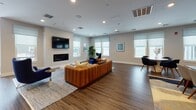 Located at 381 Court St in Plymouth, Massachusetts, this contemporary single-floor community space offers a thoughtfully designed gathering environment with a coastal-inspired aesthetic. The open-plan layout is anchored by a spacious lounge area featuring rich hardwood-style flooring, recessed lighting, and neutral gray walls that create a clean, modern backdrop throughout.

The main living area is appointed with a large leather sofa, accent chairs, and a linear gas fireplace beneath a wall-mounted flat-screen television, providing a comfortable and inviting focal point. Multiple windows dressed with soft Roman shades allow abundant natural light to fill the room while framing scenic views of the surrounding waterfront landscape.

A dedicated dining nook accommodates a round table with sleek upholstered chairs, positioned to take full advantage of the panoramic water views through oversized windows. Adjacent to the main gathering area, a well-appointed kitchenette features bold navy cabinetry, crisp white quartz countertops, a subway tile backsplash, stainless steel appliances including a dishwasher and beverage refrigerator, and a large breakfast bar with leather counter stools — ideal for casual entertaining. Open shelving adds both functionality and style. The overall ambiance is polished, welcoming, and distinctly coastal in character.