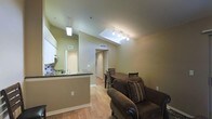 This single-floor residence showcases an open-concept layout that seamlessly connects the living, dining, and kitchen areas into one cohesive, well-lit space. The most striking architectural feature is the vaulted ceiling, which soars overhead and incorporates a skylight that floods the main living area with natural light. A track lighting system further enhances the brightness of the space, complementing the warm, neutral tones of the sage-green and beige walls throughout.

Warm-toned hardwood flooring runs continuously across the entire living area, lending the space a sense of unity and warmth. The kitchen is defined by a peninsula counter with a dark stone countertop, white upper cabinetry, and a single-basin sink — all neatly separated from the living area while maintaining an open visual connection. A pendant light hangs above the kitchen area, adding a subtle design detail.

The dining area accommodates a wood dining table with ladder-back chairs, positioned conveniently between the kitchen and the living space. The living area features a plush upholstered armchair and area rug, creating a comfortable and inviting atmosphere. A hallway visible from the main area leads toward additional rooms, including what appears to be a bedroom with bi-fold closet doors, completing this functional and well-designed single-level home.