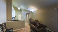This single-floor residence showcases an open-concept layout that seamlessly connects the living, dining, and kitchen areas into one cohesive, well-lit space. The most striking architectural feature is the vaulted ceiling, which soars overhead and incorporates a skylight that floods the main living area with natural light. A track lighting system further enhances the brightness of the space, complementing the warm, neutral tones of the sage-green and beige walls throughout.

Warm-toned hardwood flooring runs continuously across the entire living area, lending the space a sense of unity and warmth. The kitchen is defined by a peninsula counter with a dark stone countertop, white upper cabinetry, and a single-basin sink — all neatly separated from the living area while maintaining an open visual connection. A pendant light hangs above the kitchen area, adding a subtle design detail.

The dining area accommodates a wood dining table with ladder-back chairs, positioned conveniently between the kitchen and the living space. The living area features a plush upholstered armchair and area rug, creating a comfortable and inviting atmosphere. A hallway visible from the main area leads toward additional rooms, including what appears to be a bedroom with bi-fold closet doors, completing this functional and well-designed single-level home.