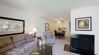This single-floor residence offers an open-concept layout that seamlessly connects the main living areas, creating a cohesive and functional living environment. The living room serves as the centerpiece of the space, featuring plush gray seating including a sofa and armchair, accented by decorative throw pillows and a dark coffee table. A fireplace adds a welcoming focal point to the room, while a framed mirror enhances the sense of openness. Neutral-toned carpet and light-colored walls contribute to a bright, airy ambiance throughout. Adjacent to the living area, the dining space accommodates a dark wood table and matching chairs, ideal for everyday meals and gatherings. A ceiling fan overhead serves both the dining and kitchen areas, ensuring comfort and air circulation. The kitchen is efficiently laid out with white cabinetry, light countertops, and standard appliances, offering a clean and practical cooking space. The overall design emphasizes an easy flow between rooms, making the unit feel spacious and well-connected. The consistent neutral color palette and coordinated furnishings create a cohesive, move-in-ready aesthetic throughout the home.