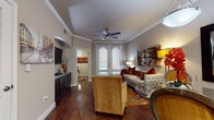 This single-floor residence is thoughtfully designed with a cohesive aesthetic that blends warm tones, quality finishes, and functional living spaces throughout. The living room serves as the heart of the home, showcasing rich hardwood flooring, elegant crown molding, and a ceiling fan. French doors fill the space with natural light and provide access to an outdoor balcony. A wall-mounted television and a sleek media console anchor one side of the room, while a tufted sofa, plush armchair, and a vibrant floral area rug create an inviting atmosphere. The open-concept layout flows seamlessly into the dining area and kitchen, where dark espresso cabinetry, stainless steel appliances, a tile backsplash, and granite countertops make a striking statement. A curved kitchen island with bar seating adds a social element to the space. A dedicated home office features hardwood floors, a glass-topped desk, and a window that provides natural light, making it ideal for focused work. The primary bedroom is generously proportioned with plush carpeting, crown molding, recessed lighting, and ample natural light. It connects to a walk-in closet and a well-designed bathroom complete with a granite-topped vanity, tile flooring, and a shower with curtained surround.