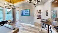 This contemporary clubhouse combines functionality with aesthetic appeal. The space showcases a harmonious blend of modern and rustic elements, featuring wood-paneled accent walls and ceilings alongside sleek furnishings. Globe-shaped chandeliers provide ambient lighting throughout, complementing the natural light from expansive windows.

The main lounge area boasts comfortable seating arrangements and a wall-mounted TV, ideal for social gatherings. A separate dining space includes a round table with distinctive blue chairs, perfect for casual meals or meetings. Art pieces adorning the walls add visual interest and character to the space.

A standout feature is the game room, complete with a pool table, which offers a prime view of the outdoor pool area through floor-to-ceiling windows. The room's wood-planked ceiling adds warmth to the space, while the pool's azure waters create a serene backdrop.

Throughout the clubhouse, the flooring alternates between warm wood tones and neutral tiles, unifying the various areas while maintaining distinct zones. The overall design emphasizes open spaces and smooth transitions, creating a welcoming environment for residents to relax, socialize, and enjoy recreational activities.