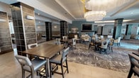 This single-floor amenity space is thoughtfully designed to serve as a premier social hub, blending contemporary style with functional comfort throughout. The main lounge area anchors the space with a striking stone-clad fireplace wall topped by a large flat-screen TV, surrounded by plush cream-upholstered seating and patterned area rugs that add warmth and texture underfoot. Bold, dark-tiled columns punctuate the open floor plan, creating natural zones for gathering while maintaining a sense of openness. A mix of high-top bar tables and standard dining sets provides flexible seating arrangements across the lounge, all illuminated by elegant layered lighting including a standout multi-tiered pendant chandelier. The fully equipped demonstration kitchen features dark cabinetry, professional-grade stainless steel appliances, a commercial range hood, and a generous quartz-topped island with bar seating beneath a trio of sculptural pendant lights — ideal for cooking events and casual entertaining. Adjacent to the lounge, a sunlit dining area opens directly to an outdoor pool deck through floor-to-ceiling sliding glass doors framed by sheer gold-patterned drapes, flooding the space with natural light and resort-style views. A private meeting room with barn-style doors and its own TV rounds out this well-appointed community space.