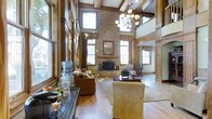 This impressive living space showcases exceptional craftsmanship and architectural detail throughout. The great room commands attention with its soaring double-height ceilings, adorned with decorative wood beams and an elegant chandelier that anchors the space. Rich, dark wood trim and millwork frame the walls, lending a warm, traditional character to the interior.

The stacked stone fireplace serves as the room's focal point, rising dramatically toward the ceiling and featuring a classic wood mantel with an ornate mirror above. Flanking the fireplace, tall arched windows allow generous natural light to filter into the space, while drapery adds a refined finishing touch.

Hardwood floors in a light, wide-plank finish run throughout, complementing the warm tones of the wood paneling and trim. Comfortable seating, including plush sofas and accent chairs, creates an inviting conversation area centered on a traditional coffee table.

Additional windows line the perimeter walls, ensuring the space remains bright and connected to the outdoors. An adjacent area, partially visible through a broad opening, suggests a well-connected and flowing floor plan ideal for both everyday living and entertaining.