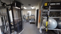 This single-floor fitness center is thoughtfully designed to support a wide range of workout routines. The space is organized into distinct functional zones, allowing users to move seamlessly between cardio, strength training, and functional fitness activities.

The cardio area is outfitted with multiple treadmills, elliptical trainers, and a stationary bike, all positioned near large windows that invite natural light and provide outdoor views, creating an energizing atmosphere during workouts. The rubber-textured flooring throughout provides durability and safety underfoot.

A Matrix cable machine anchors the strength training section, complemented by a well-stocked rack of hex dumbbells in graduated weights, paired with a weight bench and floor-to-ceiling mirrors that enhance the sense of space. The functional training zone features a dedicated wall-mounted Training Zone rack stocked with kettlebells, stability balls, resistance bands, and TRX suspension trainers, offering versatile options for bodyweight and functional exercises.

The facility is well-lit with overhead fluorescent lighting and features multiple access points, including a push-bar emergency exit door. Neutral-toned walls and clean finishes give the space a practical yet welcoming character. Additional storage closets line one wall, keeping the workout floor clear and organized. Security is supported by ceiling-mounted cameras throughout.