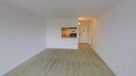 This single-floor space is designed with a straightforward, functional layout that maximizes the use of its open floor plan. The living area is finished with wall-to-wall carpeting in a neutral beige tone, contributing to a calm and understated aesthetic throughout. White walls keep the space bright and versatile, providing a clean backdrop adaptable to various interior styles.

A defining architectural feature is the pass-through window connecting the living area to the kitchen, allowing visual openness between the two spaces while maintaining a degree of separation. The kitchen is compact yet practical, equipped with a gas range and a sink positioned beneath a ventilation hood. A window in the kitchen brings in a measure of natural light, enhancing the functionality of the cooking space.

The entryway is accessible through a solid door and is finished with hard flooring, creating a practical transition zone between the exterior and the carpeted living area. Recessed and ceiling-mounted lighting provides consistent illumination throughout. Overall, the space presents a clean, move-in-ready environment with a simple and efficient design suited for everyday residential use.