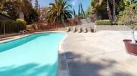 Located at 5253 El Cerrito Dr in Riverside, California, this Canyon Crest Views community amenity space offers a well-appointed outdoor retreat designed for relaxation and recreation. The centerpiece is a generously sized swimming pool with clear, blue water, framed by a broad concrete pool deck lined with lounge chairs — ideal for soaking up the Southern California sun. Mature palm trees, cypress, and colorful bougainvillea surround the area, providing natural shade and vibrant visual interest against the tiered hillside landscape.

The outdoor space extends beyond the pool area into a multi-level concrete patio, where additional seating areas are furnished with umbrella-shaded tables and chairs, creating comfortable spots for outdoor dining or socializing. Potted plants and manicured hedgerows along retaining walls add a polished, maintained aesthetic throughout.

The property's exterior architecture features neutral-toned wood siding, pitched shingle roofing, and an exterior staircase connecting levels, reflecting a classic residential style. A gated entry corridor provides secure access to the building's interior. The overall ambiance is that of a serene, community-oriented outdoor environment, well-suited to the warm, sunny climate of Riverside, CA.
