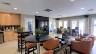This single-floor communal space is thoughtfully designed to serve multiple functions within an open, welcoming layout. The room features a prominent dark-framed entertainment wall with a large flat-screen television as its focal point, flanked by French doors that invite natural light and provide views of the outdoors, contributing to a bright and airy atmosphere.

The seating arrangement is varied and purposeful. High-top bistro tables paired with striped bar stools occupy one area, while low lounge chairs and sofas with warm orange upholstery create a more relaxed gathering zone. Dark wood tones used throughout the furniture provide a cohesive, contemporary aesthetic against the neutral beige and gray walls.

Recessed lighting is installed throughout the textured ceiling, supplementing the natural light that flows in through multiple windows and glass doors framed with decorative curtains. Potted greenery adds organic warmth to the space.

Visible through interior glass windows is an adjoining area that appears to house fitness equipment and computer workstations, suggesting this facility offers residents or guests a well-rounded set of amenities within a connected, efficiently organized floor plan.