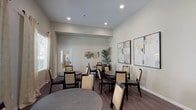 Located in the Porter Ranch neighborhood of Los Angeles, this single-floor communal dining space offers a comfortable and well-appointed environment designed for social gatherings and shared meals. The room features rich wood-look flooring that runs throughout, complementing the soft, neutral gray-toned walls and creating a warm, cohesive aesthetic.

Multiple round dining tables are arranged throughout the space, each accompanied by a set of cushioned chairs with woven backs and dark wood frames, offering ample seating for groups. Large windows dressed with elegant, patterned curtains and horizontal blinds line one side of the room, flooding the interior with generous natural light and offering views of the surrounding landscaped greenery outside.

Recessed ceiling lighting provides consistent illumination throughout, enhancing the bright and open atmosphere of the space. Decorative touches include a pair of framed abstract artworks mounted on the main wall, featuring muted gray and gold tones that complement the room's color palette. Tall potted tropical plants are strategically placed in the corners, adding a vibrant natural element to the interior.

A doorway connects this space to an adjacent room, providing functional flow between areas. Overall, the space strikes a balance between functional design and welcoming ambiance, making it well-suited for communal dining and social interaction.