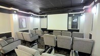 This single-floor screening room is designed to deliver a dedicated cinematic experience in a refined, lounge-style setting. The space is anchored by a large projection screen mounted at the front wall, providing a commanding focal point for all seated guests. Rows of generously sized, upholstered armchairs in a neutral gray fabric are arranged in a theater-style layout, offering comfortable seating for a sizeable group while maintaining clear sightlines to the screen.

The interior design employs a bold contrast of dark and light tones: dark-painted walls and a drop ceiling with exposed ductwork create a moody, immersive atmosphere, while a white horizontal band along the upper walls adds a clean architectural accent. Recessed and wall-mounted speakers are positioned strategically throughout the room, suggesting a built-in audio system suited for true cinematic sound.

Framed artwork on the side walls adds a touch of personality, reinforcing the room's entertainment-oriented character. A mirrored wall element near the entrance reflects light and adds visual depth to the space. Overall, the room combines functional theater design with comfortable, stylish furnishings to create an inviting private viewing environment.