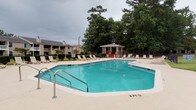 Located at 700 Martin Creel Blvd in Summerville, South Carolina, this single-story community amenity space serves as a central gathering hub for residents. The standout feature of the property is its generously sized outdoor swimming pool, which offers clear, turquoise water and a surrounding concrete deck lined with lounge chairs, ideal for relaxation and recreation. The pool area is enclosed by a black iron fence and bordered by neatly trimmed hedges and towering mature trees that provide natural shade and a sense of privacy.

A charming red-and-white gazebo with steps is positioned adjacent to the pool, offering a shaded retreat within the outdoor space. On the opposite side, a covered porch area with a blue awning, large windows, and columned supports provides direct views of the pool and surrounding landscape. This structure appears to function as a clubhouse or community gathering space, with its blue entry door and windowed façade creating an inviting, open feel.

The grounds include well-maintained green lawns with pathway lighting and decorative terra cotta planters adding warmth to the exterior. An American flag is visible near the entrance, contributing to the welcoming character of the community. The overall ambiance is relaxed, well-kept, and community-oriented.