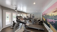 Located at 29843 Clearbrook Cir in Hayward, California, this single-room fitness center offers a well-equipped exercise space designed to support a variety of workout routines. The room is finished with neutral gray walls, dark resilient flooring suited for high-traffic use, and a textured ceiling fitted with recessed lighting and a ceiling fan to maintain comfortable airflow during workouts.

The exercise room is outfitted with multiple treadmills and elliptical trainers for cardiovascular training, along with a multi-function cable weight machine, a flat weight bench, kettlebells, and a stationary recumbent bike — providing options for both cardio and strength conditioning. A standout decorative feature is a large, vivid mural of the San Francisco Bay Bridge at sunset, adding a striking visual focal point to the space.

Natural light filters in through windows and glass-panel doors that open to an outdoor area, offering views of the surrounding grounds and lending an open, airy quality to the room. The direct outdoor access also connects the fitness center to what appears to be a pool area, enhancing the overall amenity experience. Safety and informational signage is posted throughout, and a restroom is accessible directly from the exercise room, adding practical convenience for users.