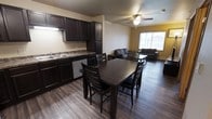This 1-bedroom, 1-bathroom unit offers a practical and comfortable open-concept floor plan designed to maximize its efficient footprint. The kitchen is outfitted with rich, dark espresso cabinetry, granite-style countertops, a stainless steel sink, and a dishwasher, providing ample storage and prep space. Under-cabinet lighting adds functional illumination to the workspace. The kitchen flows seamlessly into the dining area, which comfortably accommodates a full-sized dining table with seating for six, making it well-suited for both everyday meals and entertaining. Durable wood-look flooring runs throughout the main living areas, lending a cohesive and easy-to-maintain aesthetic. The living room is open and inviting, featuring a ceiling fan with light fixture and a window that brings in natural light while offering exterior views. The warm yellow accent wall adds a touch of color and character to the space. The overall layout is thoughtfully designed to balance the kitchen, dining, and living functions within a single, connected space, creating a comfortable and functional home environment.