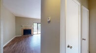 This single-floor residence at 7820 W Capitol Ave in Little Rock, Arkansas offers a well-organized layout with a comfortable blend of functional living spaces. The living room serves as the focal point of the home, highlighted by a tile-surround fireplace with a classic white mantel shelf and direct access to a private outdoor balcony or patio through a sliding glass door, allowing natural light to fill the space. Rich hardwood-style flooring flows throughout the main living and kitchen areas, adding warmth and visual continuity. The galley-style kitchen is efficiently designed with white cabinetry trimmed in wood accents, dark countertops, black appliances including a refrigerator, stove, and dishwasher, a stainless steel sink, and an overhead microwave. A pass-through window connects the kitchen to the main living area, enhancing the open feel. An adjacent pass-through view also reveals a secondary wet bar or utility area. The bedroom features plush carpeting, a ceiling fan with light fixture, a generously sized closet, and a large window that invites natural light. The bathroom is finished with full white tile surrounding a soaking tub and shower combination, complemented by classic chrome fixtures. A programmatic thermostat is conveniently located near the entryway for easy climate control.