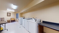 This single-floor shared facility is designed to serve the practical and recreational needs of its occupants. The space is divided into two functional areas that flow seamlessly together, finished throughout with durable light-toned flooring and warm cream-colored walls that create a clean, welcoming environment.

The laundry area is well-equipped with a row of full-sized top-loading washing machines and front-loading dryers, providing ample capacity for communal use. A built-in dark countertop with an integrated utility sink runs along one wall, offering a convenient surface for folding and sorting laundry. Overhead fluorescent lighting ensures the area is bright and practical, while a window provides natural light and ventilation.

Adjacent to the laundry section is a versatile activity and relaxation area. This room features a folding table suitable for various uses, a balance trainer and other light exercise equipment, and a comfortable seating arrangement consisting of wooden and upholstered chairs paired with a wicker side table. Framed artwork adorns the walls, adding a welcoming touch, and a window allows natural light to fill the room. An ironing board is also available, further supporting residents' daily needs.

Together, these areas reflect a thoughtfully organized communal space that balances utility with comfort.