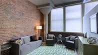 This single-floor space masterfully blends industrial heritage with contemporary design. The most striking feature is the full exposed brick wall, which lends warmth, texture, and a sense of urban history to the interior. Complementing this raw aesthetic are prominent structural columns with a smoothly finished concrete appearance, adding to the loft-like character of the space.

Large windows dressed with horizontal blinds dominate one wall, allowing generous natural light to flood the interior while offering the flexibility to control brightness and privacy. The high ceiling and open layout amplify the sense of spaciousness throughout.

The living area is thoughtfully furnished with a modern grey sofa, matching accent chairs, and a sleek glass-top side table, creating a comfortable and cohesive seating arrangement. A bold green-and-white geometric area rug anchors the space, introducing a vibrant pop of color that contrasts beautifully against the earthy brick tones. A tall floor lamp provides warm ambient lighting, enhancing the inviting atmosphere during evening hours.

Overall, the space strikes a compelling balance between the rugged charm of its industrial architectural elements and the clean lines of its contemporary furnishings, making it a distinctive and functional environment.