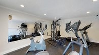 The fitness center at Woods At Killearn is a dedicated workout facility designed to support a well-rounded exercise routine. The room features a textured ceiling with recessed lighting that provides bright, even illumination throughout the space. Large floor-to-ceiling mirrors line one wall, creating a sense of openness while allowing residents to monitor their form during workouts. The space is equipped with a selection of cardio machines, including stationary bikes and an elliptical trainer, alongside a multi-station cable and weight machine ideal for full-body strength training. A treadmill is also available, rounding out the cardio offerings. The dark rubberized flooring provides both durability and comfort underfoot, making it practical for a variety of exercise types. A wall-mounted television adds convenience for entertainment during workouts. The clean, neutral color palette of white walls paired with the dark flooring gives the room a professional and functional aesthetic. This single-floor fitness amenity is thoughtfully outfitted to serve the everyday fitness needs of the community.