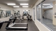 This fitness center is thoughtfully designed to provide a complete workout environment within a single, open floor layout. The space is finished with clean white walls and durable speckled rubber flooring, offering both a polished aesthetic and practical underfoot support during exercise. Overhead fluorescent lighting ensures the entire area is brightly and evenly illuminated throughout.

The cardio section is well-appointed with multiple treadmills, elliptical machines, and stationary and recumbent bikes, catering to a variety of low- and high-impact training preferences. Alongside the cardio equipment, the strength training zone features a selection of cable machines, weight benches, and resistance equipment, allowing for a comprehensive full-body regimen.

Large mirrors line the walls, enhancing the sense of openness and allowing users to monitor their form during workouts. Wall-mounted flat-screen televisions provide entertainment options for those on cardio equipment. Windows along one side of the space allow for natural light to complement the overhead lighting, creating a bright and energizing atmosphere. A dedicated exit door with clear signage ensures safe and convenient access. The overall ambiance is clean, functional, and focused—ideal for an effective fitness experience.
