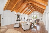 This well-appointed clubhouse offers a generously sized, single-floor layout designed for community gathering and comfort. The entrance immediately impresses with its dramatic vaulted ceiling showcasing warm, exposed wood beams that extend the full length of the space, complemented by a striking decorative floor pattern combining hardwood and tile inlays. Tall arched windows flood the foyer with abundant natural light, highlighting the crisp white wainscoting and paneled walls throughout.

The open lounge area transitions seamlessly from the entrance, featuring the same soaring cathedral ceiling with richly toned wooden beams and pendant lighting. This communal living space is anchored by a stone-accented fireplace with a wall-mounted television, making it an inviting focal point. French doors with glass panes provide access to additional areas while maintaining a bright, airy atmosphere. Comfortable seating arrangements are thoughtfully positioned throughout, offering views to the outdoors through large windows.

The kitchen is practical and neatly designed, featuring granite countertops, grey cabinetry, a double basin sink, and full-size appliances including a refrigerator and microwave — well-suited to support events and gatherings. The consistent use of light hardwood flooring, neutral tones, and coastal-inspired décor throughout the clubhouse creates a cohesive, welcoming ambiance.