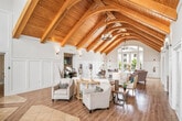 This well-appointed clubhouse offers a generously sized, single-floor layout designed for community gathering and comfort. The entrance immediately impresses with its dramatic vaulted ceiling showcasing warm, exposed wood beams that extend the full length of the space, complemented by a striking decorative floor pattern combining hardwood and tile inlays. Tall arched windows flood the foyer with abundant natural light, highlighting the crisp white wainscoting and paneled walls throughout.

The open lounge area transitions seamlessly from the entrance, featuring the same soaring cathedral ceiling with richly toned wooden beams and pendant lighting. This communal living space is anchored by a stone-accented fireplace with a wall-mounted television, making it an inviting focal point. French doors with glass panes provide access to additional areas while maintaining a bright, airy atmosphere. Comfortable seating arrangements are thoughtfully positioned throughout, offering views to the outdoors through large windows.

The kitchen is practical and neatly designed, featuring granite countertops, grey cabinetry, a double basin sink, and full-size appliances including a refrigerator and microwave — well-suited to support events and gatherings. The consistent use of light hardwood flooring, neutral tones, and coastal-inspired décor throughout the clubhouse creates a cohesive, welcoming ambiance.
