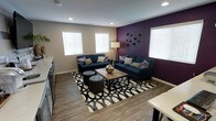 This single-story space centers around a well-appointed living area that blends contemporary design with warm, inviting elements. The room is defined by a striking deep purple accent wall, which serves as a bold focal point and adds depth and personality to the overall aesthetic. Neutral taupe walls on the remaining sides create a balanced contrast, ensuring the space feels both vibrant and cohesive.

The living area is furnished with two navy blue sofas adorned with coordinating accent pillows in purple and tan tones, arranged around a rustic wooden coffee table. A black-and-white geometric area rug anchors the seating arrangement, adding graphic interest underfoot. Two round tufted ottomans provide additional flexible seating. A floor lamp in the corner contributes warm ambient lighting, complementing the recessed ceiling lights above.

Wood-look flooring runs throughout, offering durability and a clean, modern appearance. Multiple windows fitted with white horizontal blinds allow natural light to filter into the room from several directions. Decorative wall accents on the purple wall add an artistic finishing touch. A glimpse of a dining area is visible at the edge of the frame, suggesting an open, connected layout.