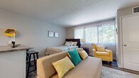 This single-floor studio unit offers a well-organized, open-concept layout that maximizes functionality. The entry hallway, accented by a welcoming sign and textured walls, transitions smoothly into the main living area, which features rich, dark wood-look flooring throughout. Large windows dressed with vertical blinds allow generous natural light to fill the space, creating an airy and inviting atmosphere.

The main living area serves as both a living room and sleeping space, comfortably accommodating a sofa, seating chair, and a full bed. A wall-mounted flat-screen television is positioned above a wicker storage cabinet, flanked by floor lamps that provide warm ambient lighting. A small breakfast bar with a granite-style countertop and barstool separates the living area from the kitchen, offering a convenient dining option.

The kitchen is equipped with stainless steel appliances, including an electric range and over-the-range microwave. White cabinetry, a subway tile backsplash, and a stainless steel sink contribute to a clean, modern aesthetic.

The bathroom features a elongated, narrow layout with white shaker-style cabinetry, a tub-shower combination with marble-look tile surround, and coordinating wood-look vinyl flooring, completing the cohesive design of the unit.