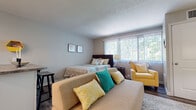 This single-floor studio unit offers a well-organized, open-concept layout that maximizes functionality. The entry hallway, accented by a welcoming sign and textured walls, transitions smoothly into the main living area, which features rich, dark wood-look flooring throughout. Large windows dressed with vertical blinds allow generous natural light to fill the space, creating an airy and inviting atmosphere.

The main living area serves as both a living room and sleeping space, comfortably accommodating a sofa, seating chair, and a full bed. A wall-mounted flat-screen television is positioned above a wicker storage cabinet, flanked by floor lamps that provide warm ambient lighting. A small breakfast bar with a granite-style countertop and barstool separates the living area from the kitchen, offering a convenient dining option.

The kitchen is equipped with stainless steel appliances, including an electric range and over-the-range microwave. White cabinetry, a subway tile backsplash, and a stainless steel sink contribute to a clean, modern aesthetic.

The bathroom features a elongated, narrow layout with white shaker-style cabinetry, a tub-shower combination with marble-look tile surround, and coordinating wood-look vinyl flooring, completing the cohesive design of the unit.