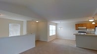 This single-floor residence offers a clean, open-concept layout that seamlessly connects the living, dining, and kitchen areas into one cohesive living space. The interior is finished in soft, neutral tones throughout, creating a calm and welcoming ambiance. The living area features wall-to-wall carpet, contributing to a warm and comfortable feel underfoot.

A white kitchen pass-through counter separates the kitchen from the main living space while maintaining an open sightline, allowing natural light and conversation to flow freely between rooms. The dining area is defined by a classic chandelier overhead, adding a subtle decorative touch to the otherwise understated space.

Natural light enters through a centrally positioned window dressed with horizontal blinds, brightening the interior during the day. The entry area is clearly defined, with a paneled door and a ceiling-mounted light fixture providing practical illumination.

White trim and baseboards frame the walls throughout, offering a crisp contrast to the beige carpeting and light-colored walls. The overall design is straightforward and functional, making it an easily adaptable space suitable for a variety of furniture arrangements and personal styles. The layout prioritizes openness and ease of movement throughout the main living areas.