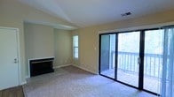 This single-floor space centers around a spacious living room finished with plush wall-to-wall carpeting and warm, neutral-toned walls that create a clean, understated ambiance. A tray ceiling adds a subtle architectural detail overhead, lending a sense of structure and dimension to the room. A fireplace with a dark surround anchors one wall, serving as a natural focal point and providing both function and visual interest. Natural light enters through a narrow vertical window on the adjacent wall, complementing the abundant light that pours in through large sliding glass doors. These doors open directly onto an outdoor deck with a white railing, where mature trees are visible beyond, offering a pleasant, leafy backdrop. The transition between the carpeted living area and the tiled threshold near the fireplace and sliding doors adds a practical layer of material contrast. The layout feels open and versatile, well-suited for a variety of furniture arrangements. The combination of the fireplace, outdoor access, and natural greenery views contributes to a comfortable, relaxed atmosphere throughout the space.