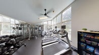 Located at 970 St Francis Blvd in Daly City, California, this fitness center is a single-level facility designed to support a comprehensive workout experience. The space is defined by its expansive floor-to-ceiling windows along one wall, which flood the interior with natural light and provide views of an outdoor pool and surrounding greenery, creating an open and motivating atmosphere.

The main exercise area features rubber flooring throughout and is equipped with a variety of cardio machines, including multiple treadmills and elliptical trainers, several of which are fitted with individual monitor screens. A multi-station cable weight machine and a dumbbell rack with accompanying bench provide dedicated strength training options along the mirrored wall, which visually expands the space.

A designated functional training corner stores stability balls in multiple sizes, exercise mats, and a vertical medicine ball rack, offering versatility for stretching and core-focused workouts. An additional area near the entrance includes a wall-mounted television, a snack and supplies cabinet, and informational displays, providing practical amenities for users. A ceiling fan aids in ventilation alongside the overhead fluorescent lighting. The glass-door entrance connects the fitness center directly to the outdoor area.